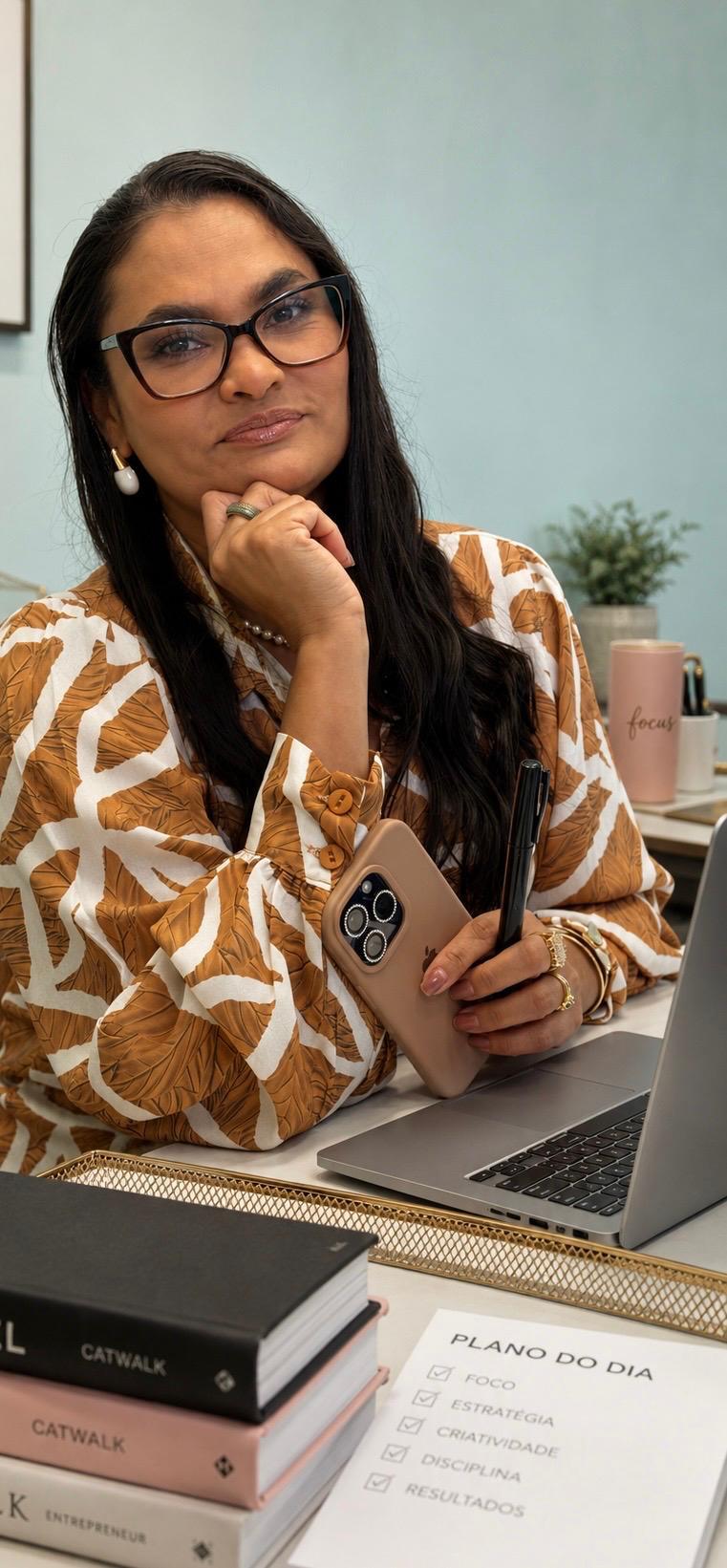 Kariny Costa, de óculos e blusa estampada em caramelo e branco, sentada à mesa de trabalho com a mão no queixo